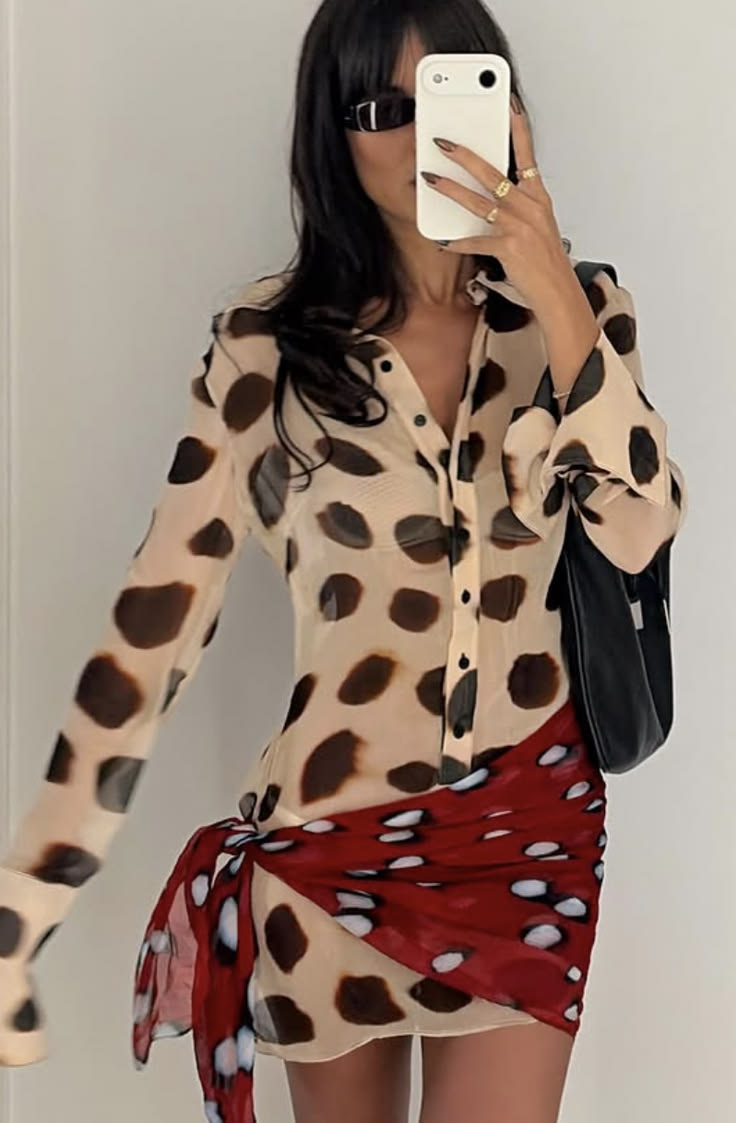 A woman takes a mirror selfie wearing a beige long-sleeve button-up shirt dress with dark brown spots, paired with a red wrap skirt featuring white spots, and carrying a black shoulder bag.