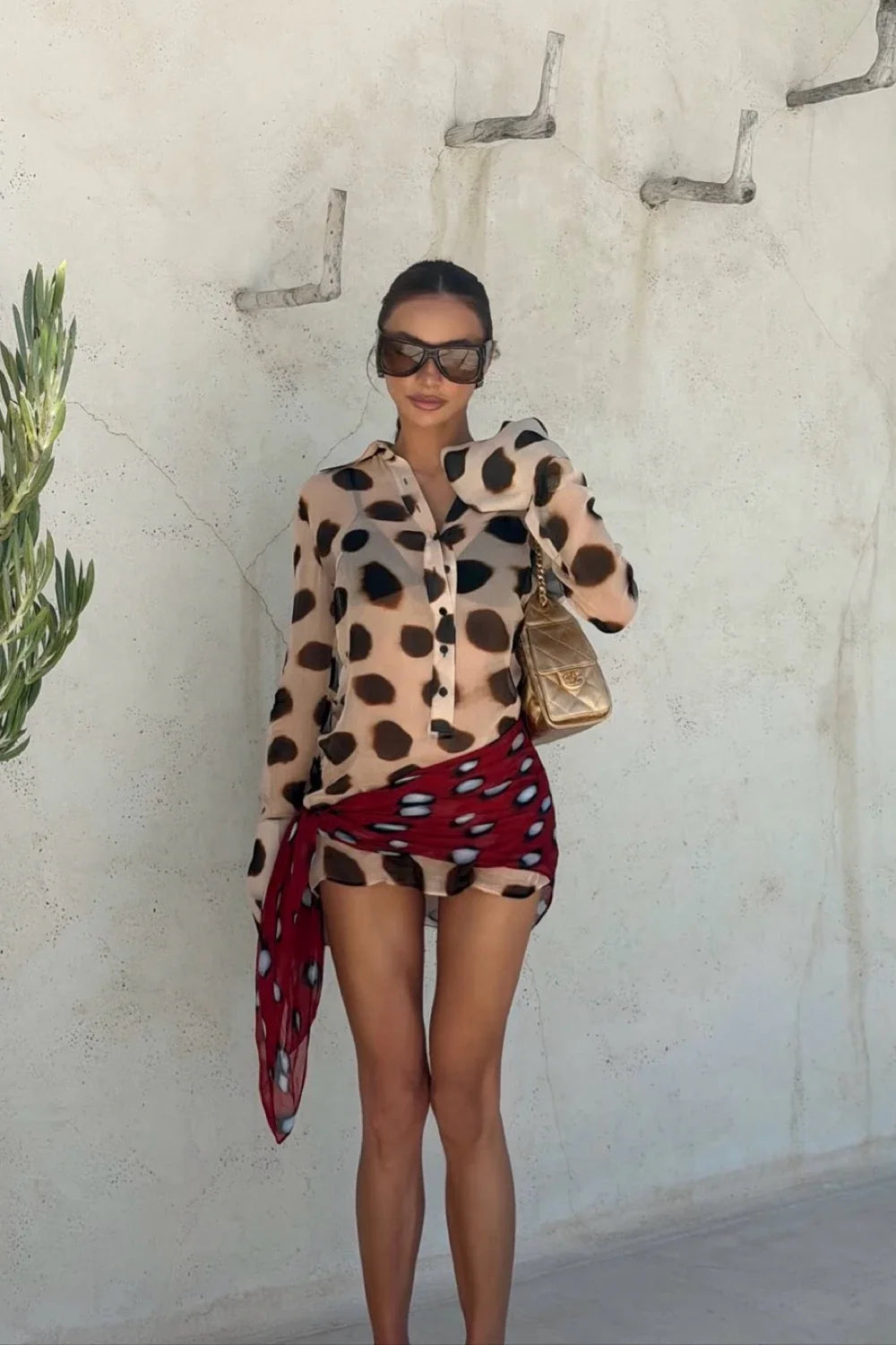 A woman stands against a textured off-white wall wearing a beige long-sleeve button-up shirt with black spots, a red wrap skirt with white spots, and sunglasses. She is holding a small gold handbag.