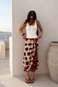 A woman wearing a white tank top and a long wrap skirt with a beige, brown, and red polka dot pattern, standing in a minimalist outdoor setting with a large stone pot and sea view in the background.