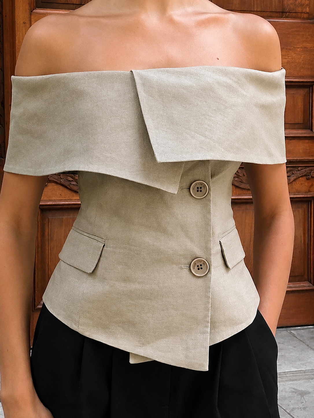 An off-the-shoulder beige linen corset top with a double-breasted button closure and flap pockets, worn by a model in front of a dark wood door.