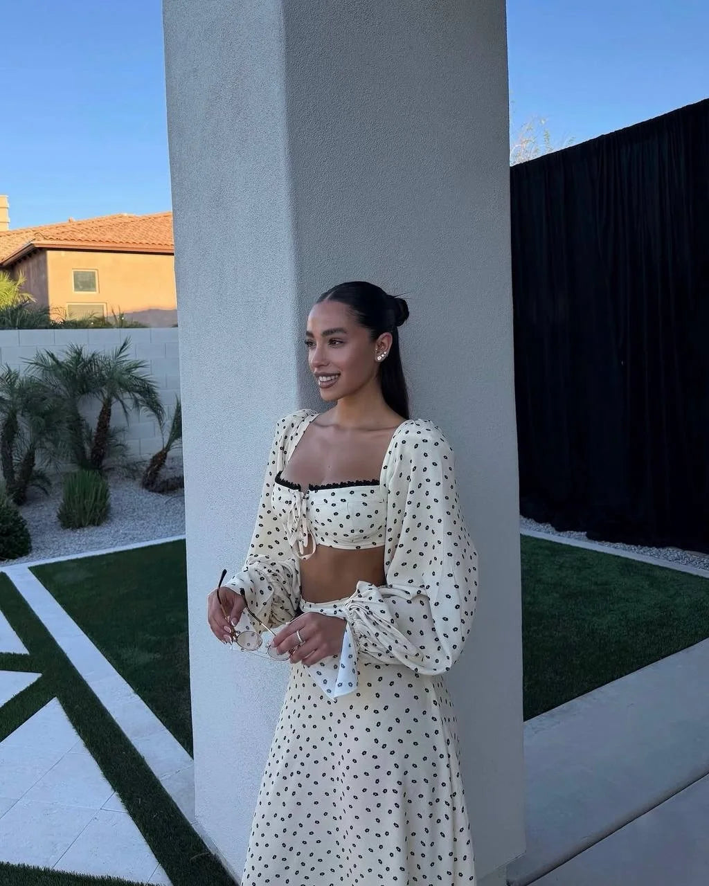 Woman in a polka dot outfit standing against a white wall with a garden and house in the background.
